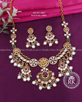 Gold-plated Indian traditional jewelry set featuring a statement necklace and matching earrings with white faux pearls, clear crystals, and green and purple stones, displayed on a purple background with a pink fabric accent.